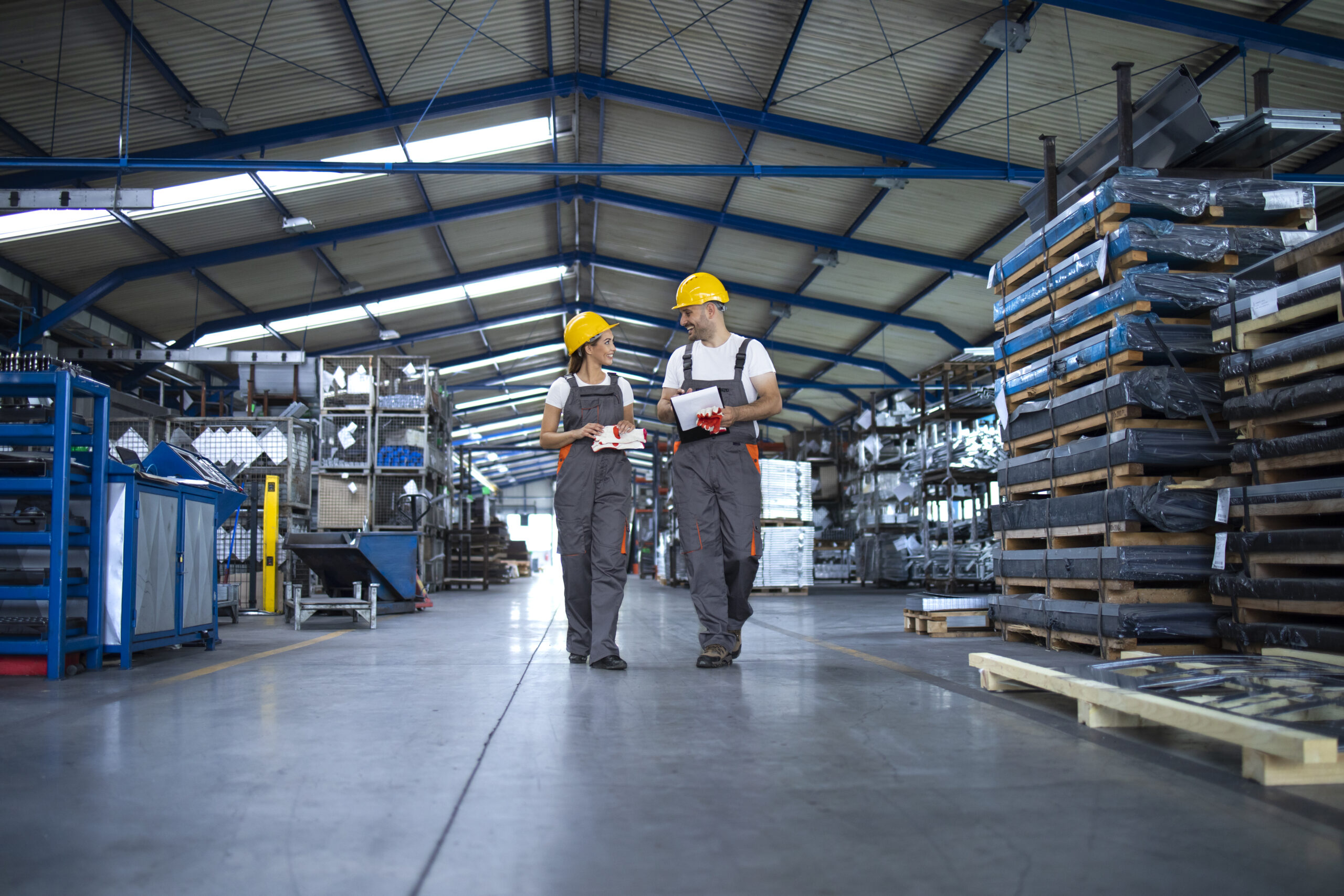 Factory workers in work wear and yellow helmets walking through industrial production hall and discussing about delivery deadline.